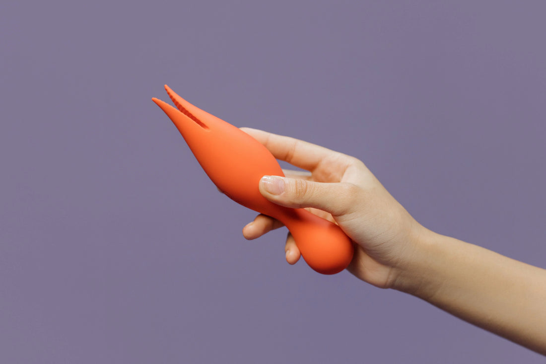 Hand holding an orange silicone clitoral vibrator with a flexible two-prong tip against a purple background