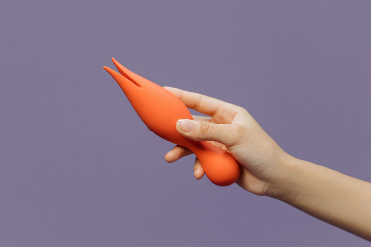 Hand holding an orange silicone clitoral vibrator with a flexible two-prong tip against a purple background