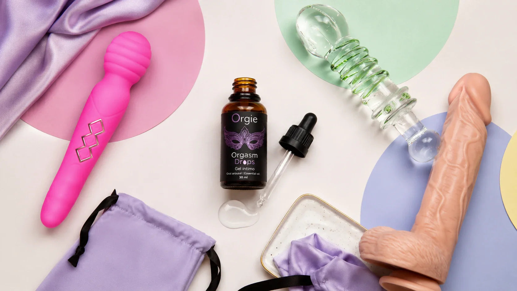 Flat lay of Orgie Orgasm Drops arousal gel for women displayed with adult wellness toys, a storage pouch, and pastel self-care styling on a clean off-white background.
