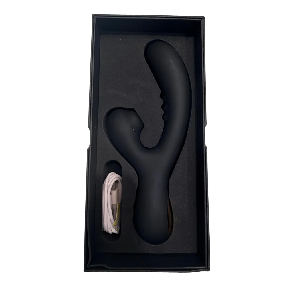 A matte black dual-stimulation personal massager shown inside its black presentation box with a foam insert, alongside a coiled white charging cable, on a plain white background.