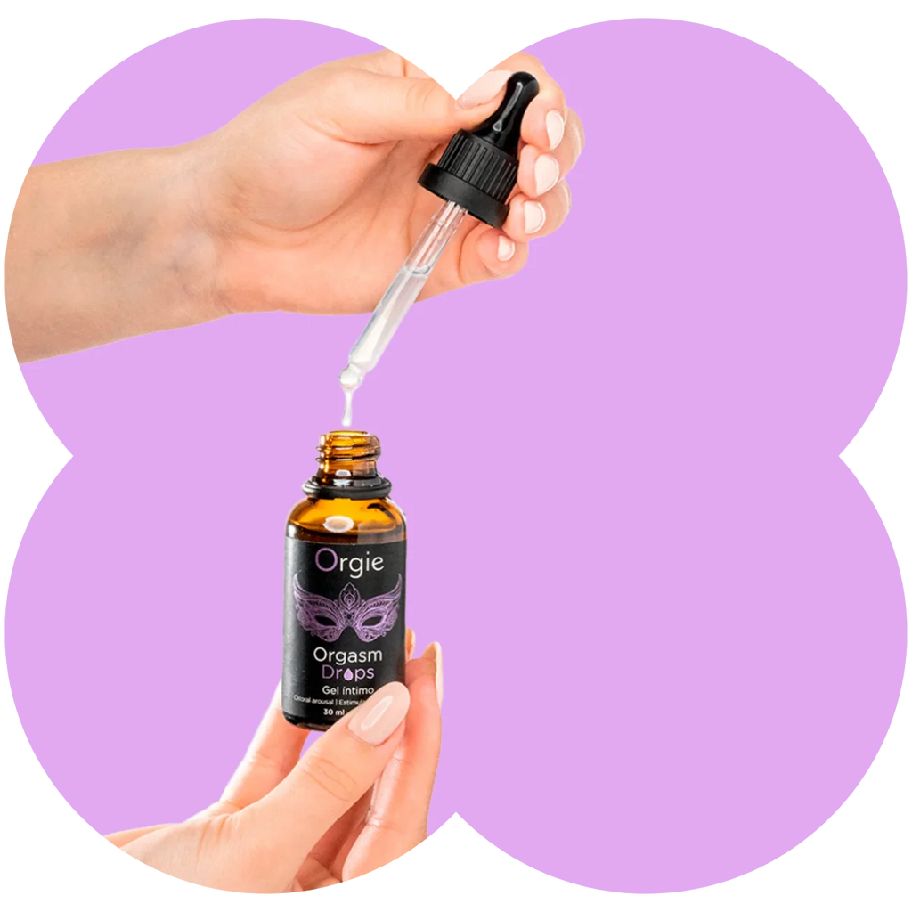 A hand holds an amber glass dropper bottle labeled Orgie Orgasm Drops while another hand positions a clear dropper above the open bottle, with a light purple abstract background.