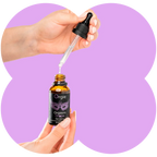 A hand holds an amber glass dropper bottle labeled Orgie Orgasm Drops while another hand positions a clear dropper above the open bottle, with a light purple abstract background.
